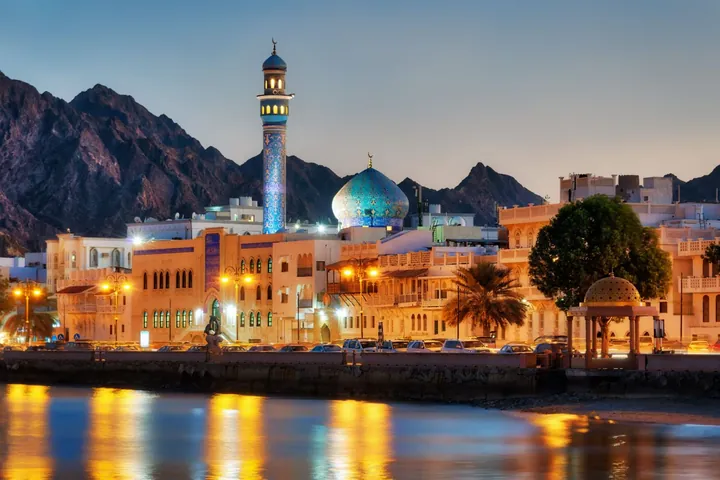A mosque dome and minaret lit at dusk on the Muscat waterfront